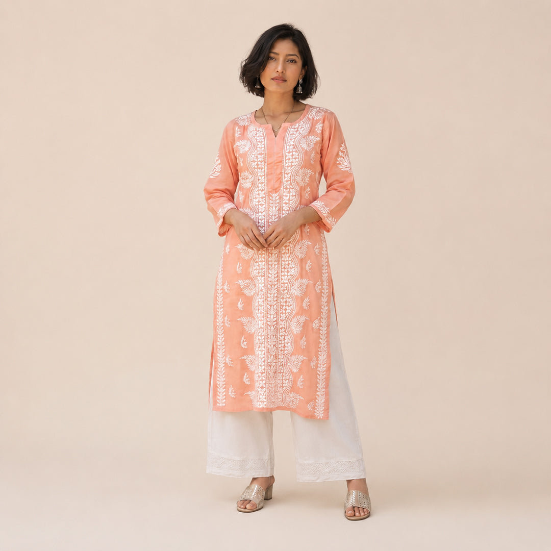 A woman modeling a peach-orange kurta with white floral embroidery and white wide-leg pants, standing against a solid beige studio background.