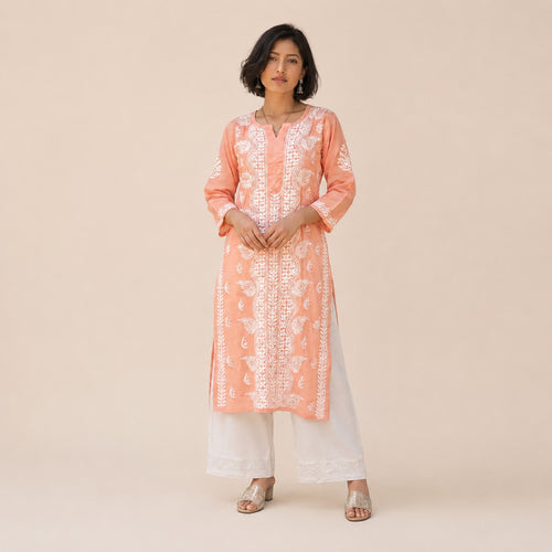 A woman modeling a peach-orange kurta with white floral embroidery and white wide-leg pants, standing against a solid beige studio background.