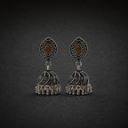 A pair of ornate silver-toned jhumka earrings with black oxidized detailing, featuring a teardrop-shaped top with a brown marquise-cut stone and a bell-shaped dangling charm with intricate filigree and small hanging beads, set against a dark textured background.