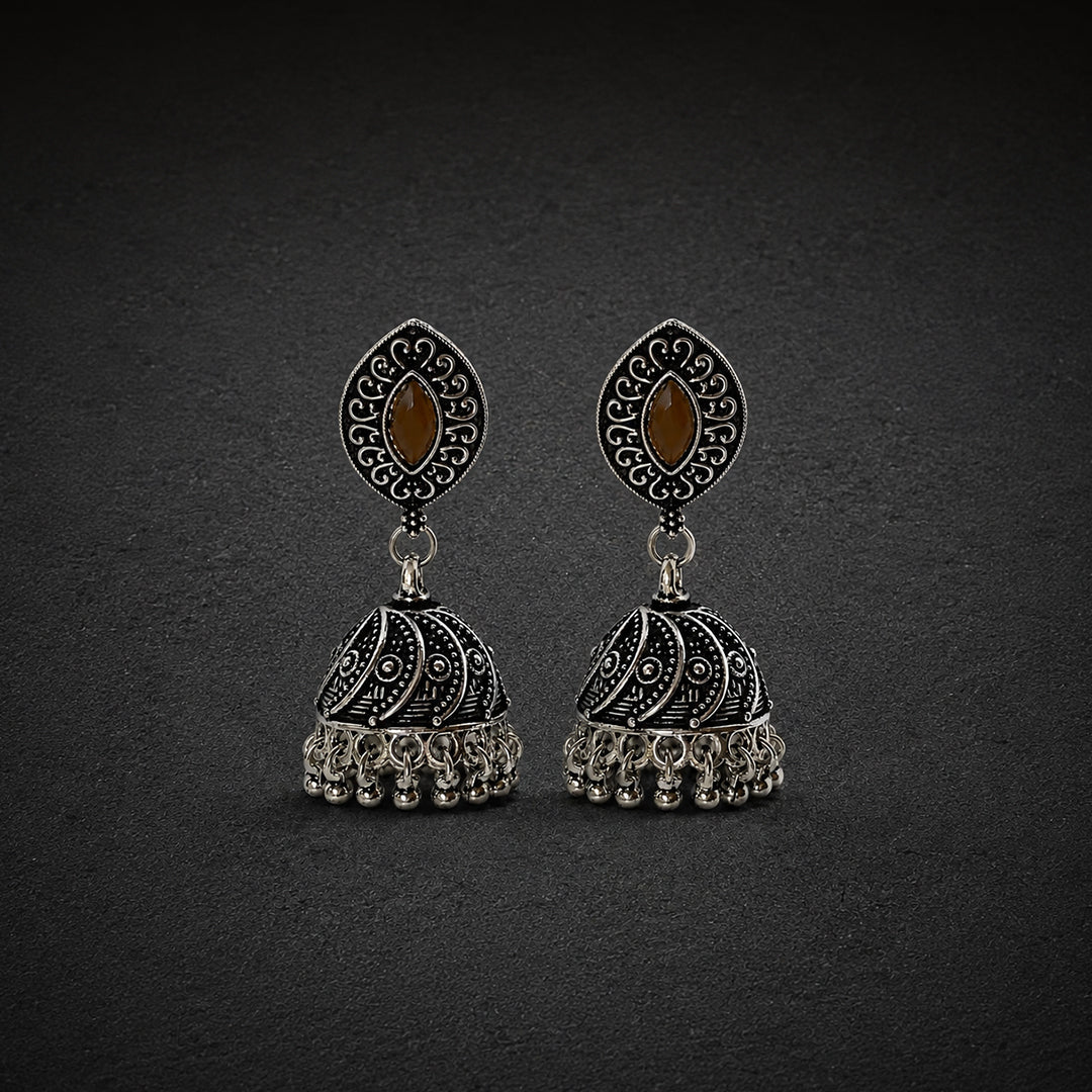 A pair of ornate silver-toned jhumka earrings with black oxidized detailing, featuring a teardrop-shaped top with a brown marquise-cut stone and a bell-shaped dangling charm with intricate filigree and small hanging beads, set against a dark textured background.