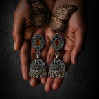 A pair of ornate silver-toned jhumka earrings with oval amber-colored stones, held in two hands with a butterfly resting above them against a dark textured background.