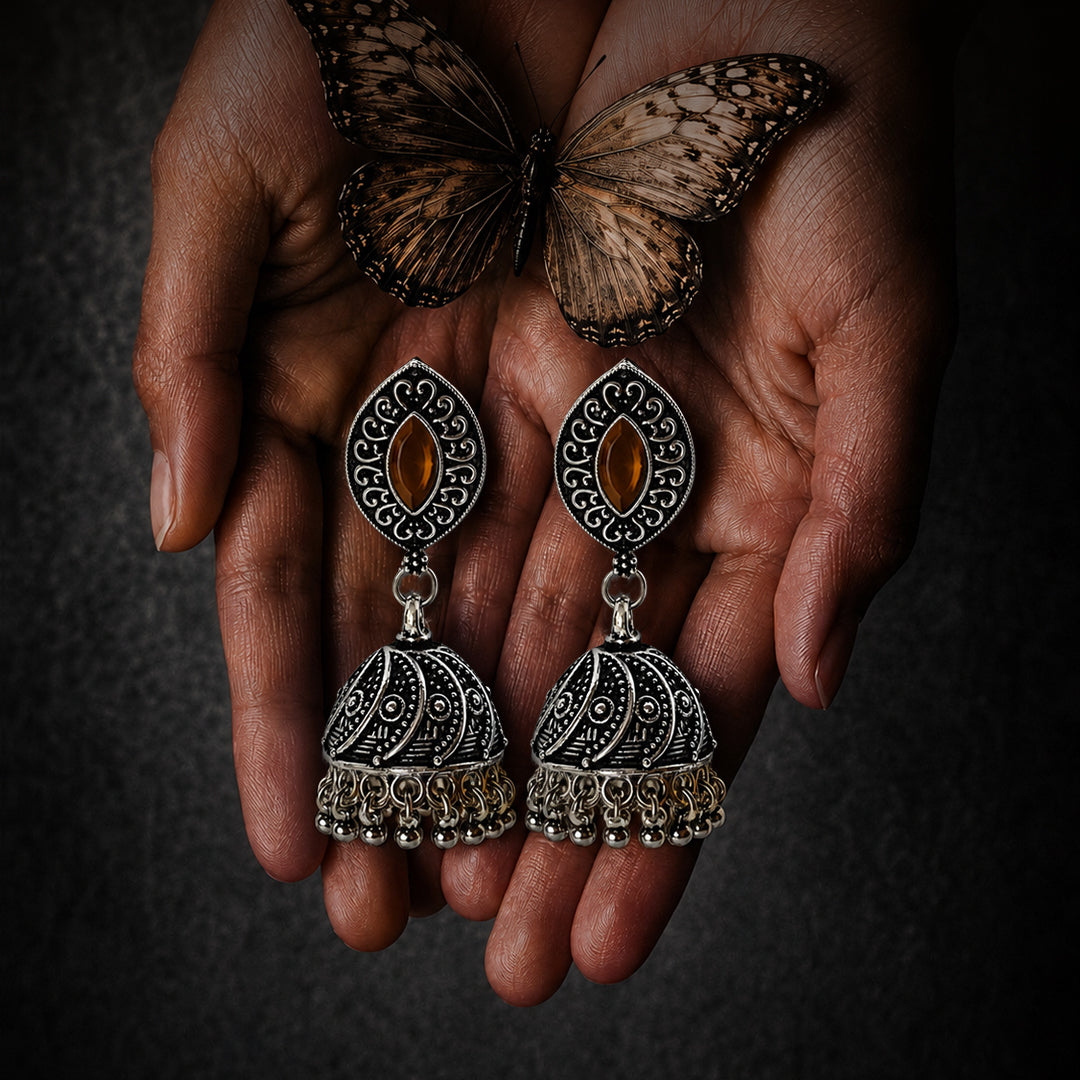 A pair of ornate silver-toned jhumka earrings with oval amber-colored stones, held in two hands with a butterfly resting above them against a dark textured background.