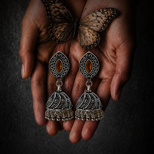A pair of ornate silver-toned jhumka earrings with oval amber-colored stones, held in two hands with a butterfly resting above them against a dark textured background.