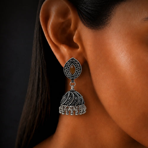 A close-up of a woman's ear wearing a silver-toned filigree jhumka earring with a teardrop-shaped top and a dangling bell-shaped charm with small hanging stones.