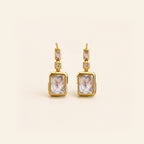 A pair of gold drop earrings featuring rectangular clear crystals in gold bezel settings, accented with small round clear stones, shown on a light cream background.