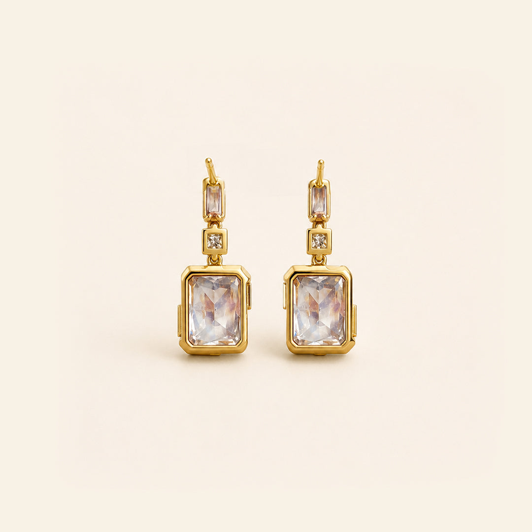 A pair of gold drop earrings featuring rectangular clear crystals in gold bezel settings, accented with small round clear stones, shown on a light cream background.