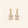 A pair of gold drop earrings featuring a large rectangular clear gemstone, a smaller rectangular stone above it, and a small square accent stone, set against a solid cream background.