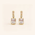 A pair of gold drop earrings featuring a large rectangular clear gemstone, a smaller rectangular stone above it, and a small square accent stone, set against a solid cream background.