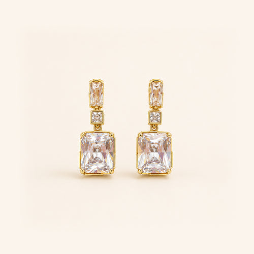 A pair of gold drop earrings featuring a large rectangular clear gemstone, a smaller rectangular stone above it, and a small square accent stone, set against a solid cream background.