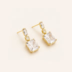 A pair of gold drop earrings featuring large clear rectangular gemstones in square prong settings, with smaller clear baguette and round-cut stones above, displayed on a light cream background.