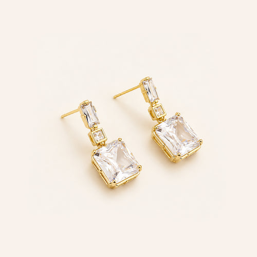 A pair of gold drop earrings featuring large clear rectangular gemstones in square prong settings, with smaller clear baguette and round-cut stones above, displayed on a light cream background.