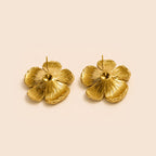 A pair of textured gold flower stud earrings on a solid pale beige background.