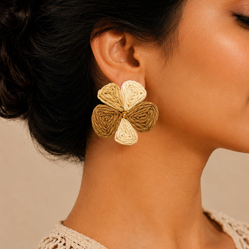 A close-up of a woman's ear wearing a large flower-shaped dangle earring made of woven raffia in natural beige and light brown tones, set against a soft beige background.