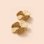 A pair of handmade braided flower earrings in shades of tan and cream, displayed on a solid pale peach background.