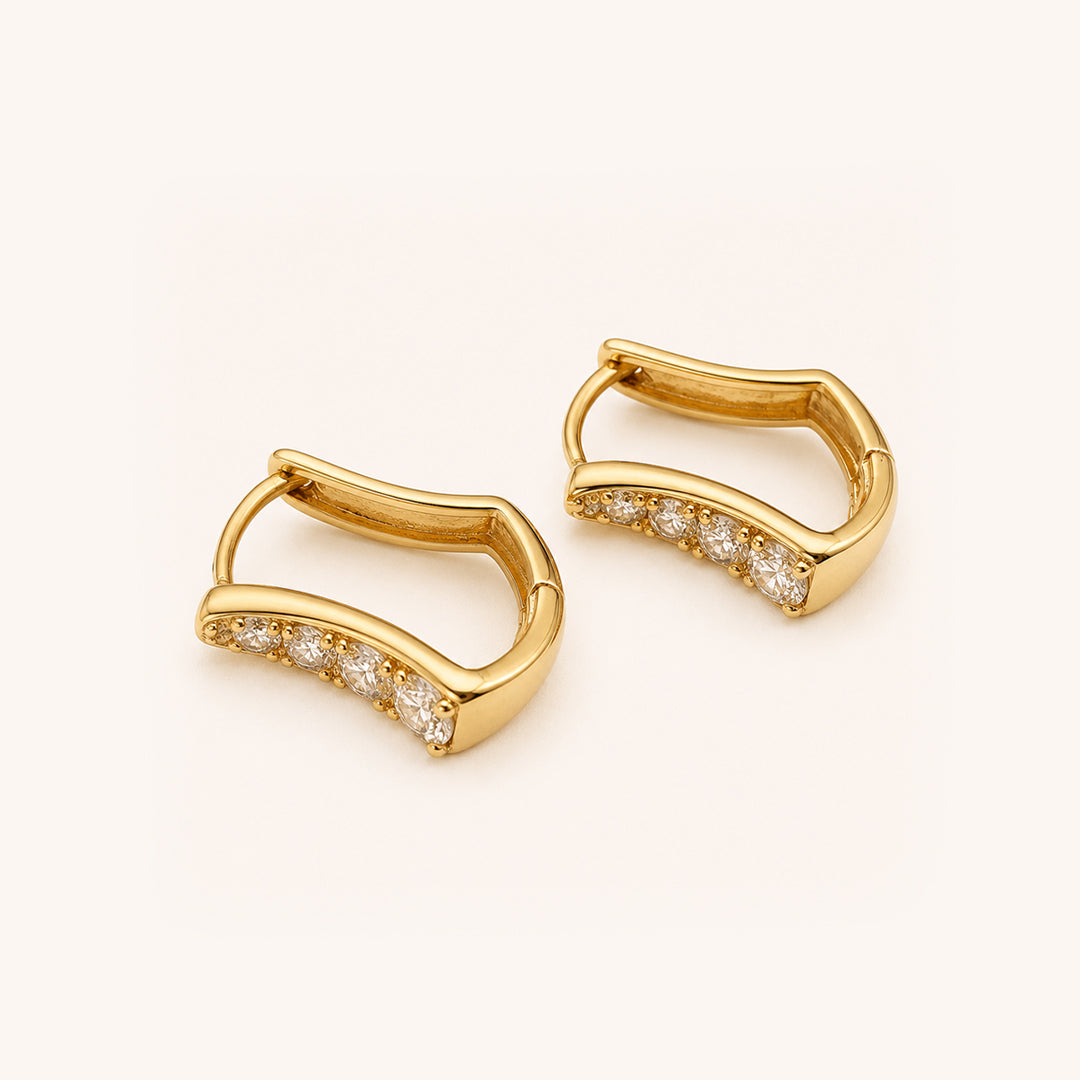 A pair of gold huggie earrings with a row of pavé-set clear stones along the front, displayed on a clean off-white background.
