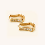 A pair of gold huggie earrings with a row of clear round-cut stones along the front, displayed on a clean off-white background.