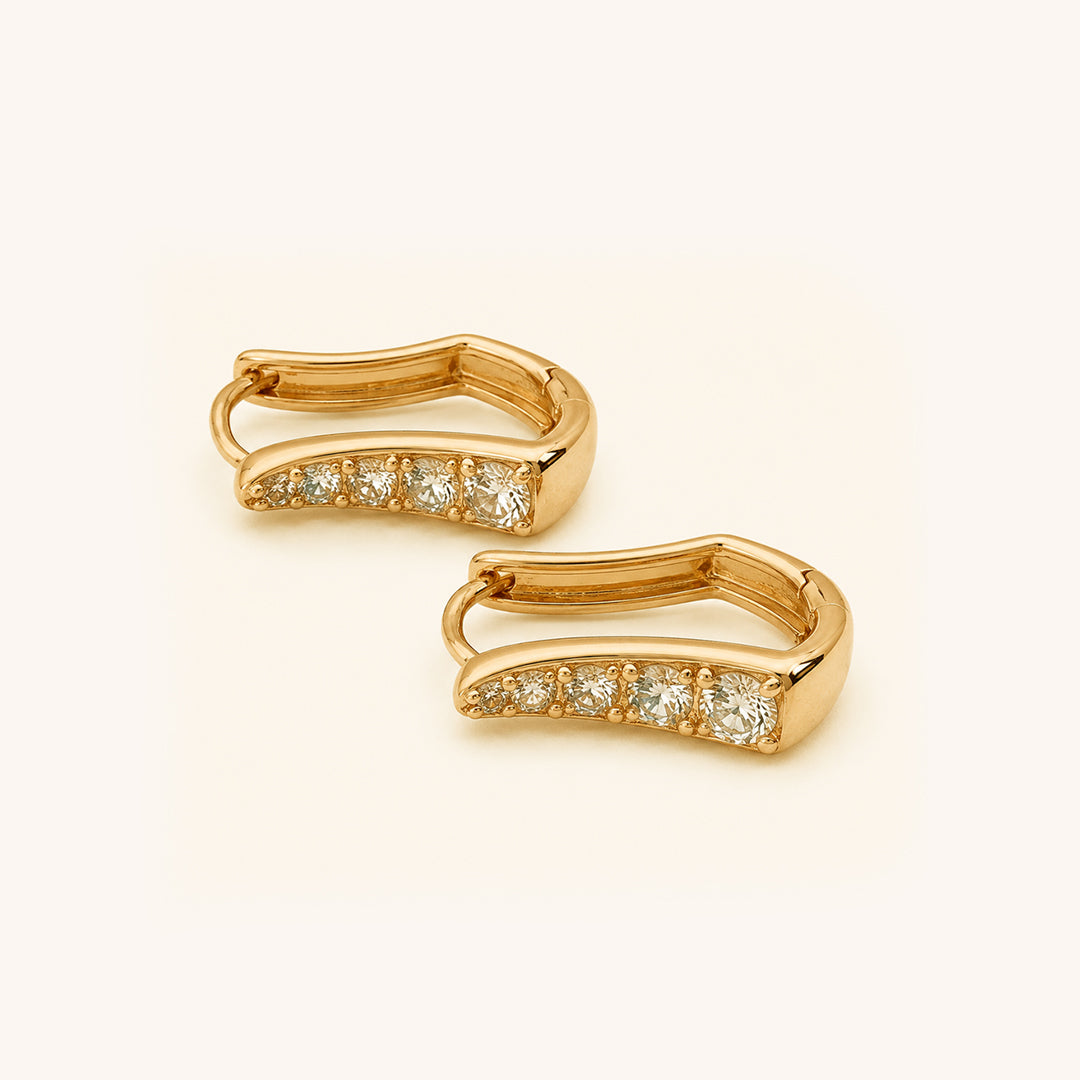 A pair of gold huggie earrings with a row of clear round-cut stones along the front, displayed on a clean off-white background.