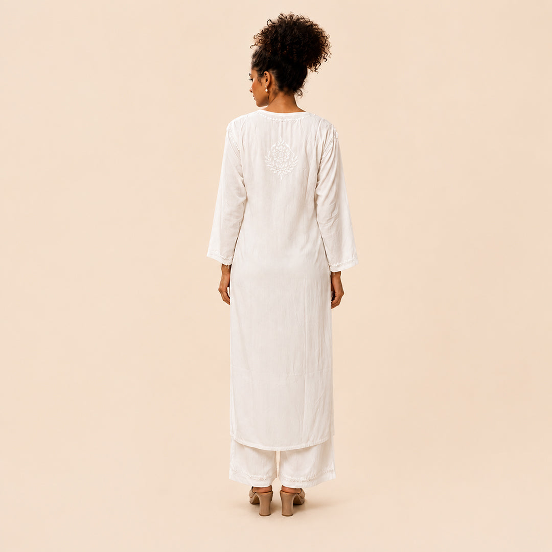 A woman viewed from the back wearing a long white tunic with three-quarter sleeves and matching wide-leg pants, standing against a plain warm beige studio background.