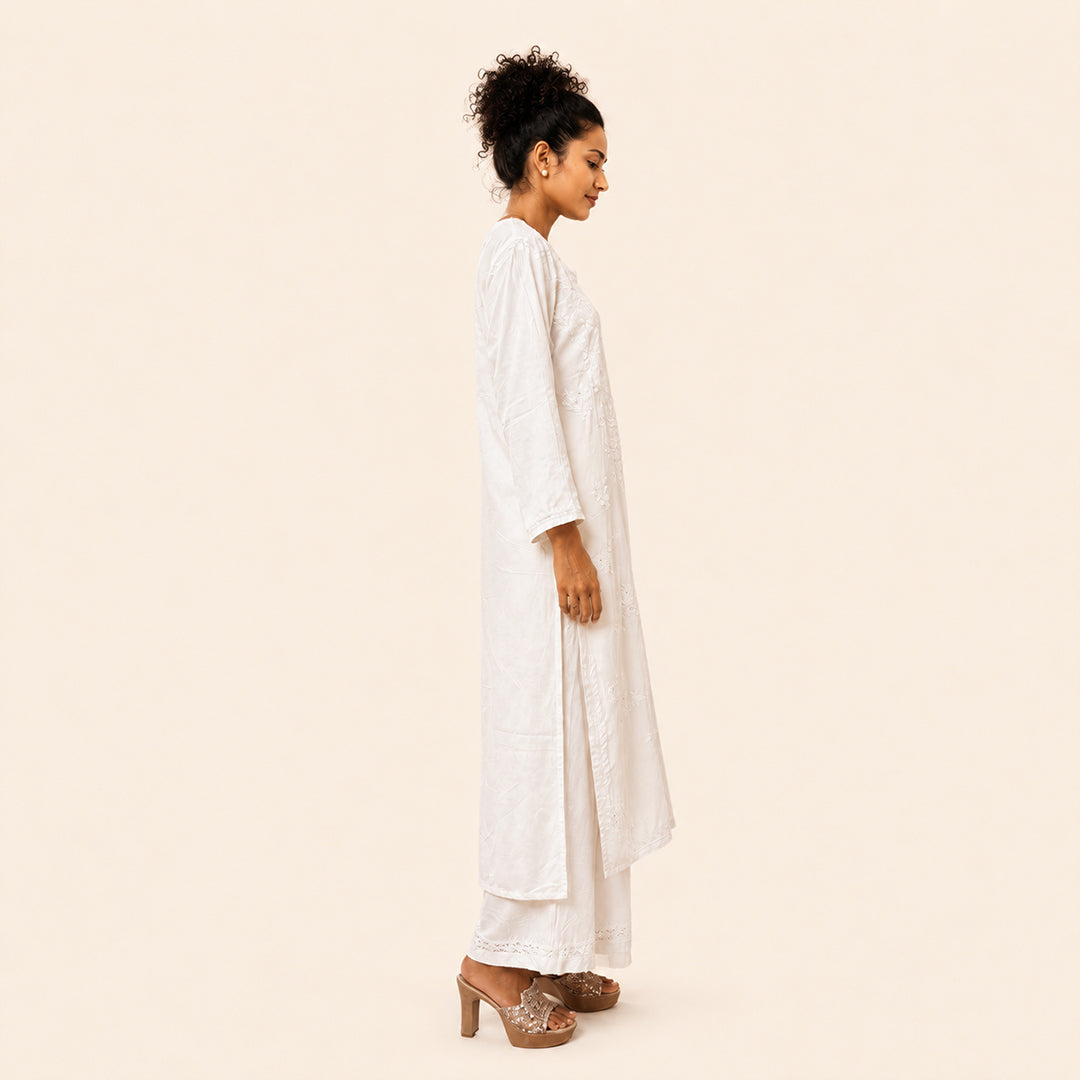 A woman shown in profile wearing a long white tunic with matching wide-leg pants and brown heeled sandals, standing against a plain warm beige studio background.