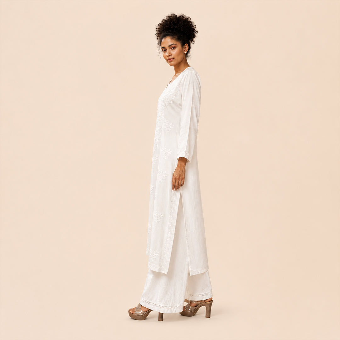A woman modeling a white long kurta with matching wide-leg pants and brown high-heeled sandals, standing in profile against a solid light beige background.