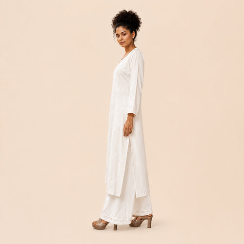 A woman modeling a white long kurta with matching wide-leg pants and brown high-heeled sandals, standing in profile against a solid light beige background.