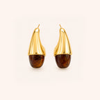 A pair of gold crescent-shaped hoop earrings with a polished amber-brown resin  drop looks like tortoshell on the end, shown against a light cream background.