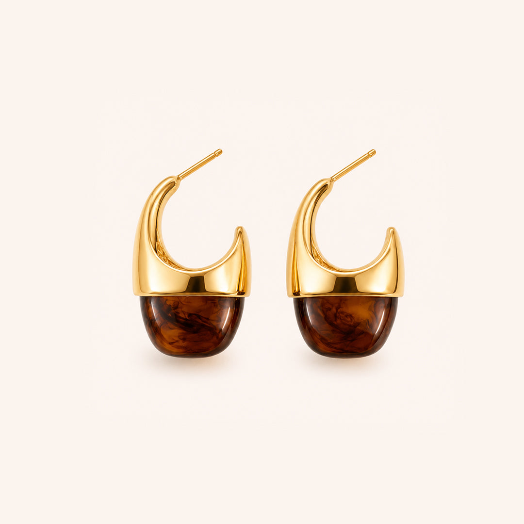 A pair of gold crescent-shaped hoop earrings with a polished amber-brown resin  drop looks like tortoshell on the end, shown against a light cream background.