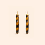 A pair of open hoop earrings with a glossy Leopard pattern in dark brown and black, framed by a polished gold-tone metal border and post, set against a solid pale peach background.