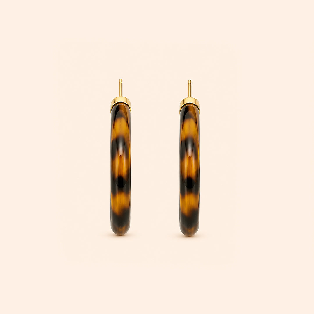 A pair of open hoop earrings with a glossy Leopard pattern in dark brown and black, framed by a polished gold-tone metal border and post, set against a solid pale peach background.