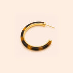 A pair of open hoop earrings with a glossy Leopard pattern in dark brown and black, framed by a polished gold-tone metal border and post, set against a solid pale peach background.