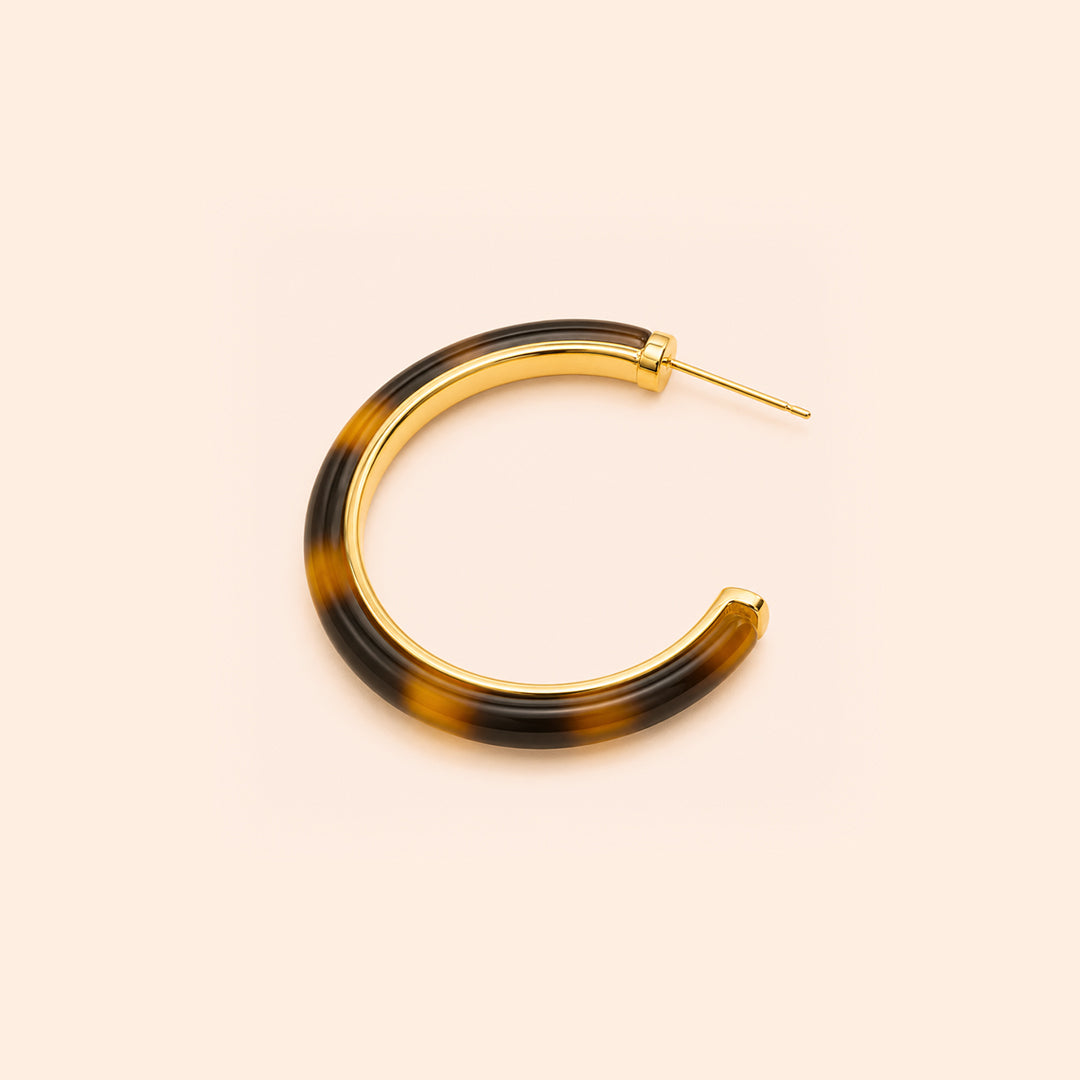 A pair of open hoop earrings with a glossy Leopard pattern in dark brown and black, framed by a polished gold-tone metal border and post, set against a solid pale peach background.