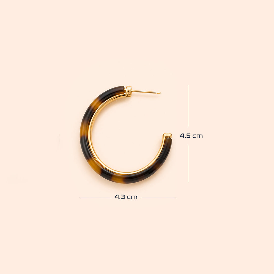 A pair of open hoop earrings with a glossy Leopard pattern in dark brown and black, framed by a polished gold-tone metal border and post, set against a solid pale peach background with size.