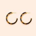 A pair of open hoop earrings with a glossy Leopard pattern in dark brown and black, framed by a polished gold-tone metal border and post, set against a solid pale peach background.