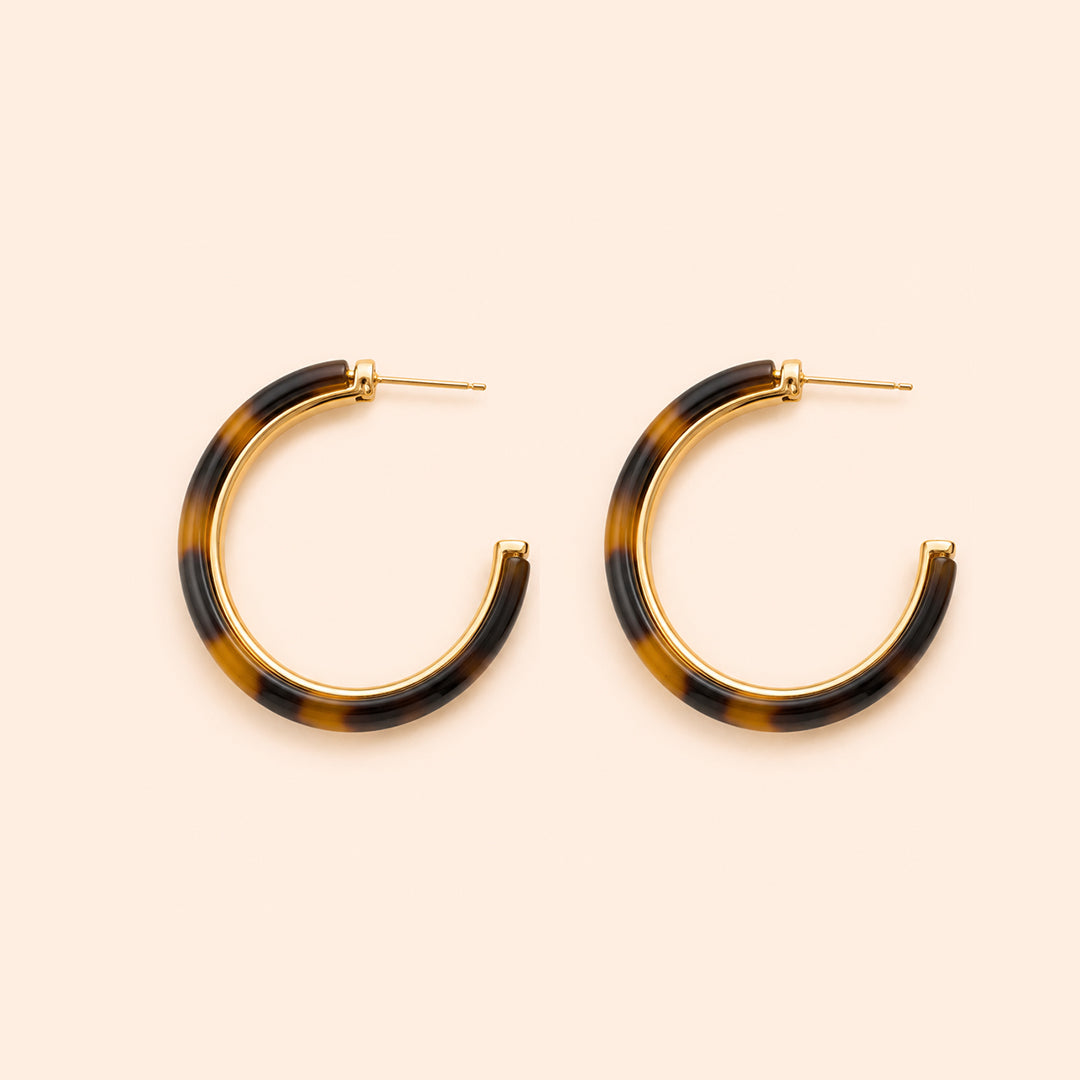 A pair of open hoop earrings with a glossy Leopard pattern in dark brown and black, framed by a polished gold-tone metal border and post, set against a solid pale peach background.