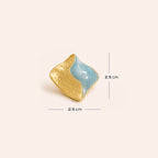 An organic-shaped gold and light blue earrings with textured gold surfaces and smooth blue inlays, displayed on a warm off-white background with size.