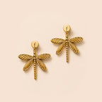 A pair of gold dragonfly stud earrings with textured wings and post backs, displayed on a solid pale peach background.