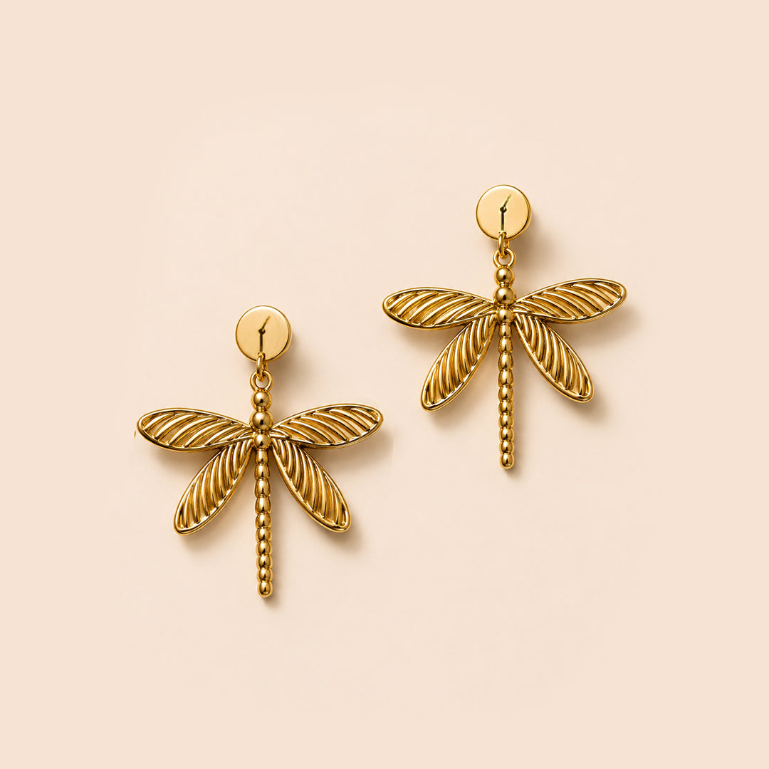 A pair of gold dragonfly stud earrings with textured wings and post backs, displayed on a solid pale peach background.