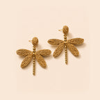 A pair of gold dragonfly earrings with textured woven wings and beaded stems, displayed on a light beige background.