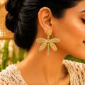 A close-up of a woman wearing a large gold dragonfly-shaped dangle earring. The earring features a textured circular stud and a body with four detailed wings, shown against the model's ear and neck.