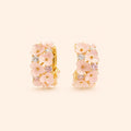 A pair of gold huggie hoop earrings decorated with pink cherry blossom flowers and small clear and pink gemstones, set against a solid pale beige background.