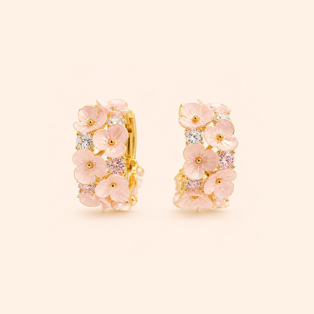 A pair of gold huggie hoop earrings decorated with pink cherry blossom flowers and small clear and pink gemstones, set against a solid pale beige background.