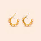 A pair of gold crescent-shaped hoop earrings decorated with light pink floral accents and small clear square crystals, shown on a solid pale peach background.