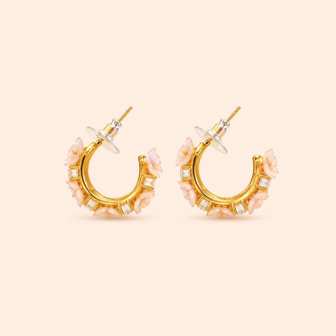 A pair of gold crescent-shaped hoop earrings decorated with light pink floral accents and small clear square crystals, shown on a solid pale peach background.
