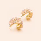 A pair of gold crescent-shaped hoop earrings decorated with pink cherry blossoms and small clear stones, set against a solid pale peach background.