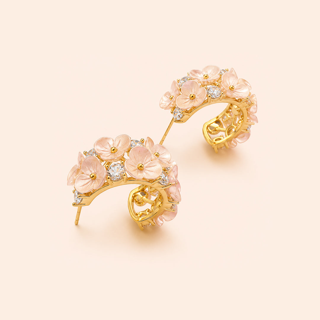 A pair of gold crescent-shaped hoop earrings decorated with pink cherry blossoms and small clear stones, set against a solid pale peach background.