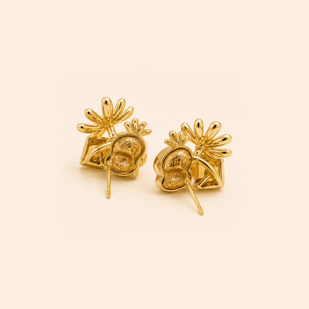 A pair of gold stud earrings with a textured, organic leaf-like design and small flower-like tops, displayed on a solid light beige background.