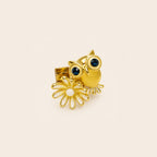 A gold owl and flower ring on a light beige background. The ring features a polished gold owl with dark blue gemstone eyes and a gold flower with a white center.