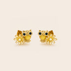 A pair of gold owl stud earrings with dark green enamel eyes, white enamel beaks, and small white flower charms, displayed on a light beige background.