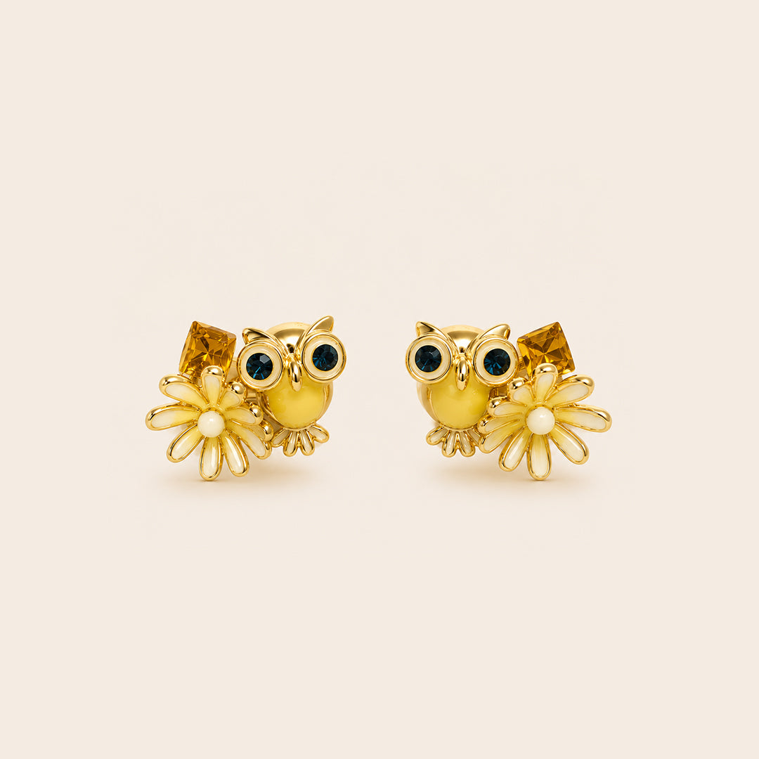 A pair of gold owl stud earrings with dark green enamel eyes, white enamel beaks, and small white flower charms, displayed on a light beige background.