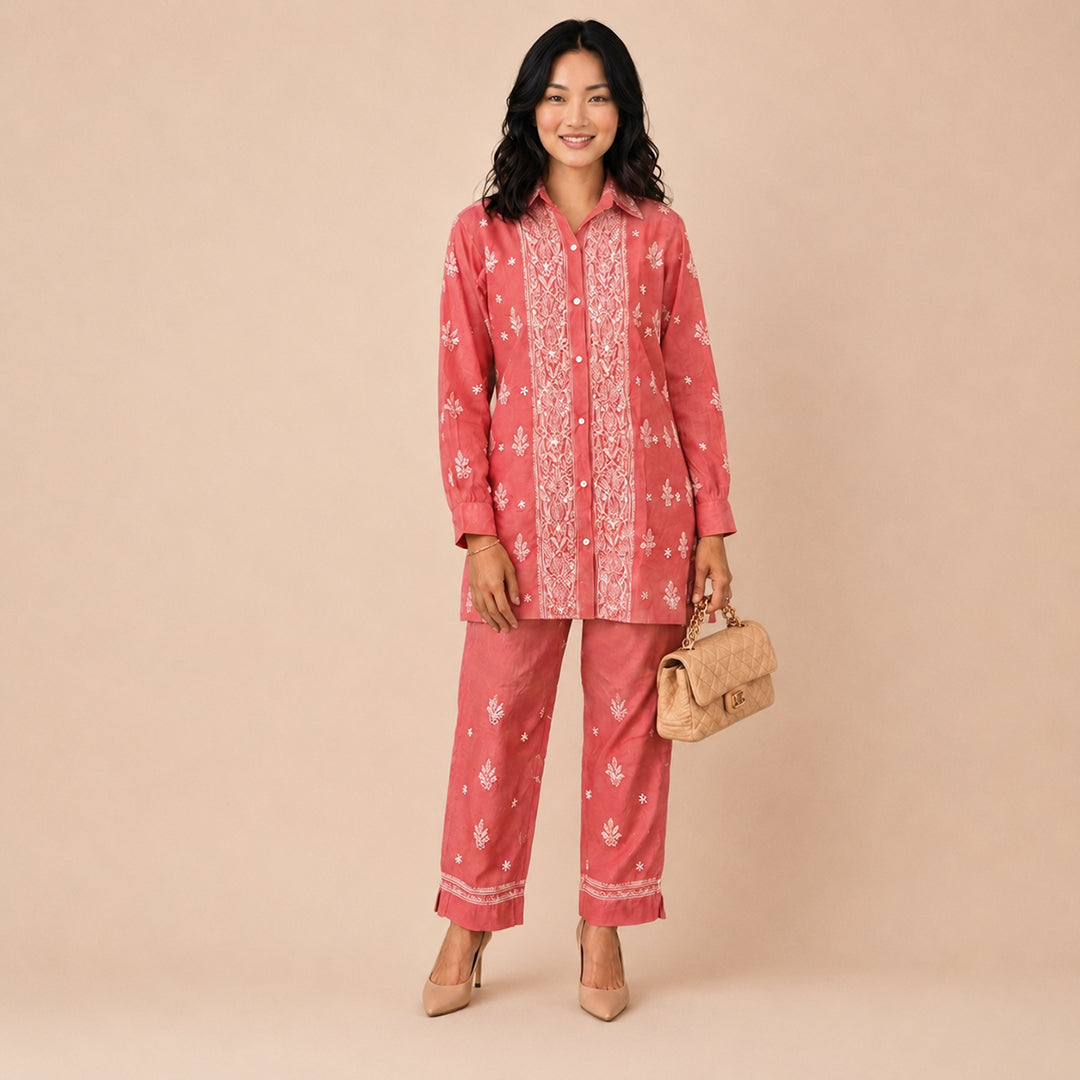 A woman modeling a two-piece pink batik pajama set with white floral patterns, consisting of a long-sleeve button-up shirt and matching pants, holding a small woven handbag against a solid beige background.